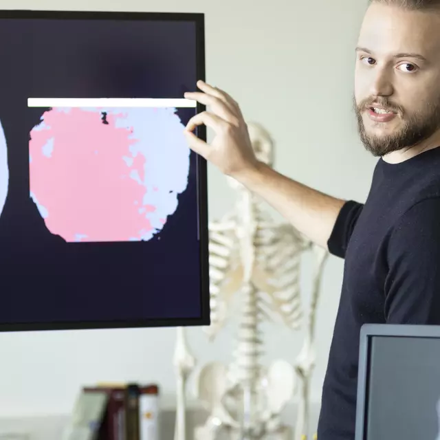 Auf einem Bildschirm sind drei mehrfarbige, kreisförmige Abbildungen abgebildet, die sich voneinander unterscheiden. Ein junger Mann steht vor dem Bildschirm und erläutert, was zu sehen ist.