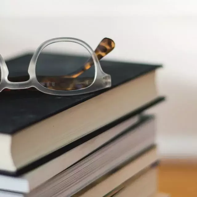 Auf einen Holztisch liegt ein Stapel Bücher. Oben drauf liegt eine Brille.