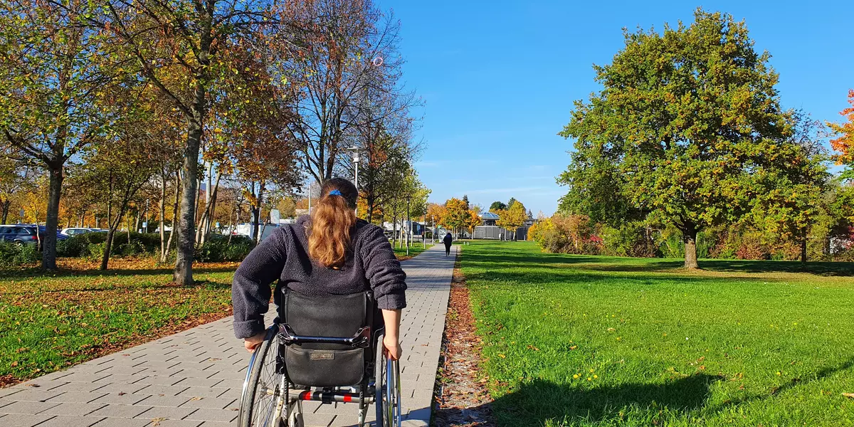 Eine Rollstuhlfahrerin auf einem Weg am OTH Campus. Rechts und links sind Bäume und eine grüne Wiese.