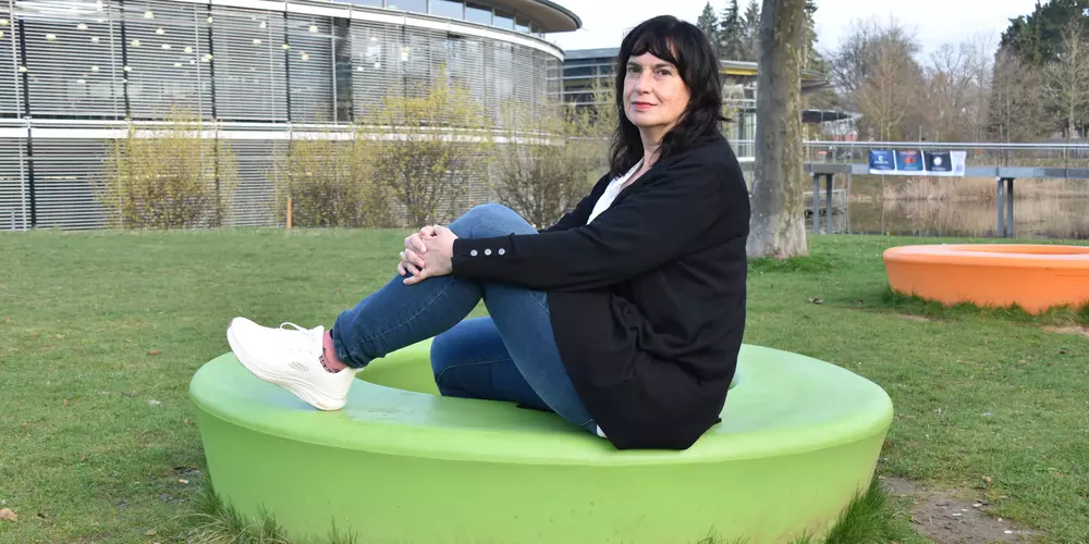 Petra Knoch sitzt auf einem grünen Sitzkreis in der Wiese. Im Hintergund das OTH Gebäude