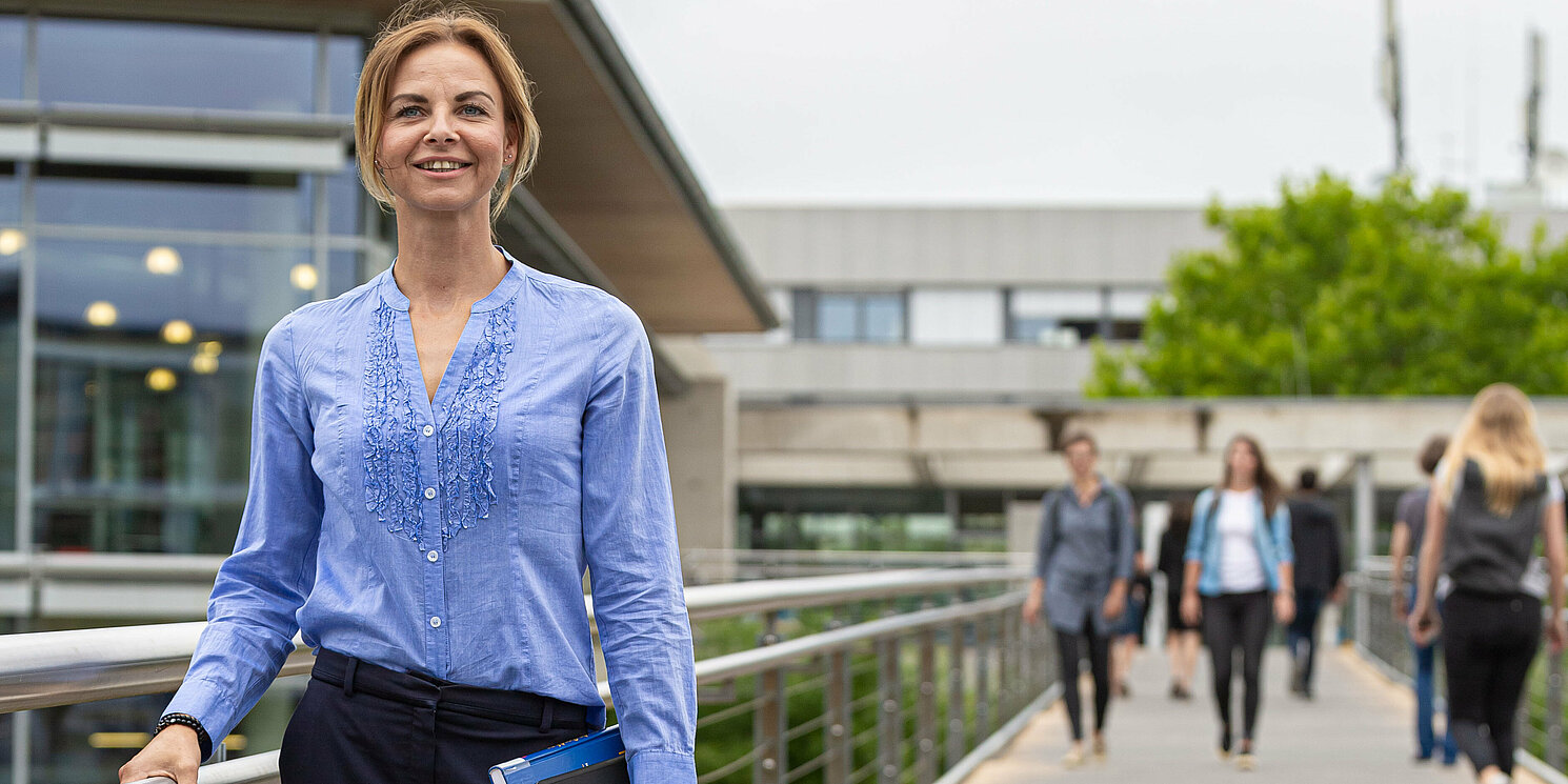 Studentin steht am Übergang zur Bibliothek. Im Hintergrund sind weitere Studierende zu sehen. 