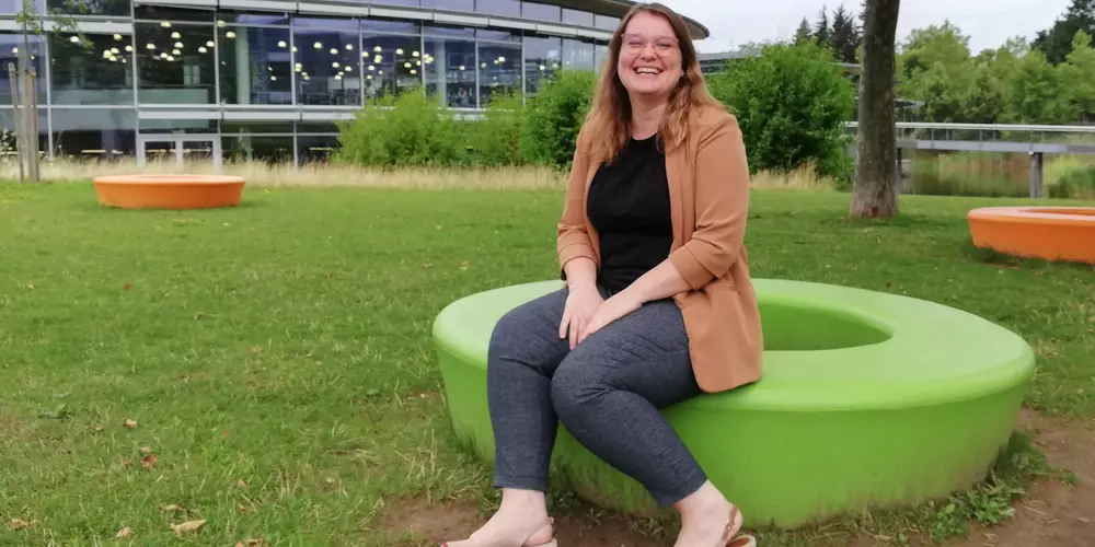 Verena Obermeier sitzt auf einem grünen Sitzkreis in der Wiese. Im Hintergund das OTH Gebäude