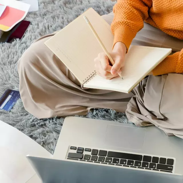 Studentin sitzt auf dem Boden auf einem grauen Teppich. Vor ihr ein Laptop, neben ihr Arbeitsblätter. Sie macht sich Notizen in ein Heft.