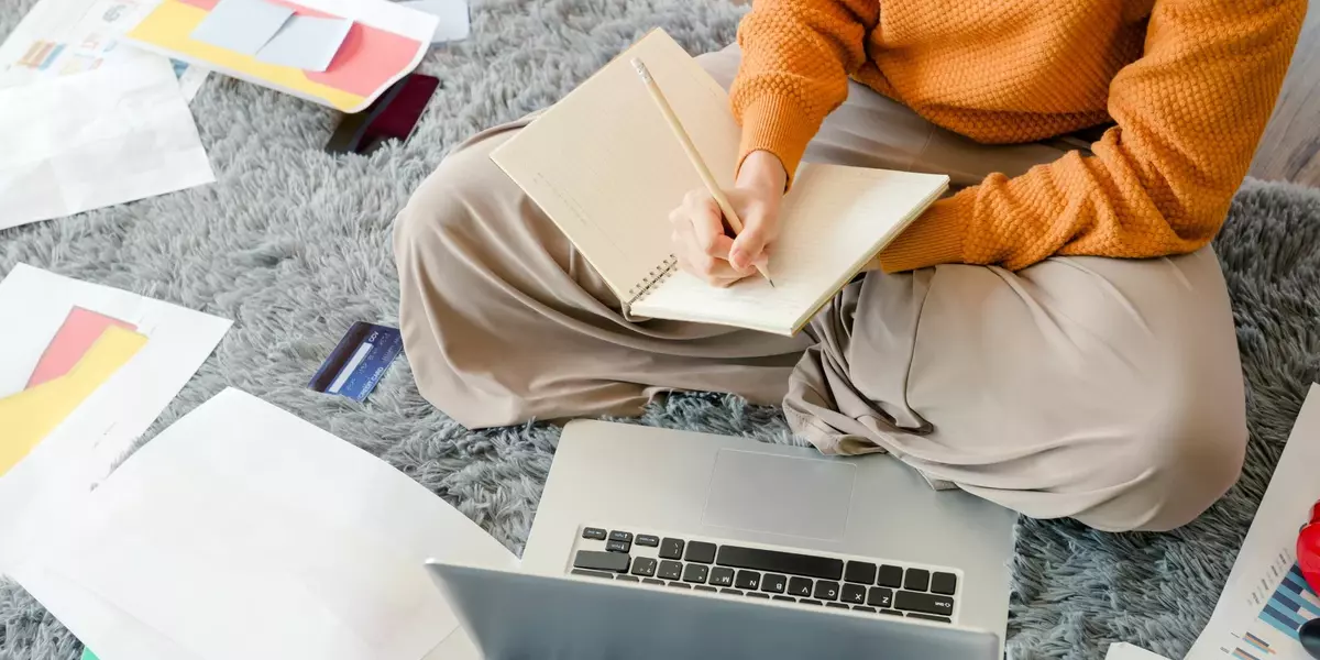 Studentin sitzt auf dem Boden auf einem grauen Teppich. Vor ihr ein Laptop, neben ihr Arbeitsblätter. Sie macht sich Notizen in ein Heft.