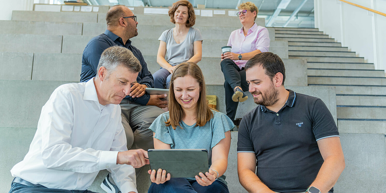 Studierende sitzen mit ihrem Dozenten auf einer großen Treppe und er erklärt ihnen etwas an einem Laptop.