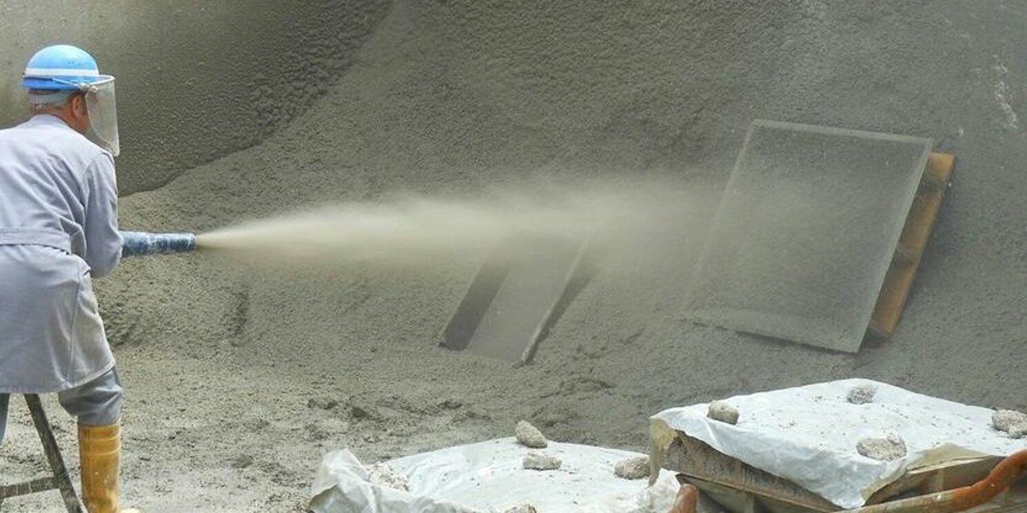 A man in protective gear holds a hose in his hand and uses it to apply shotcrete to a tunnel wall.