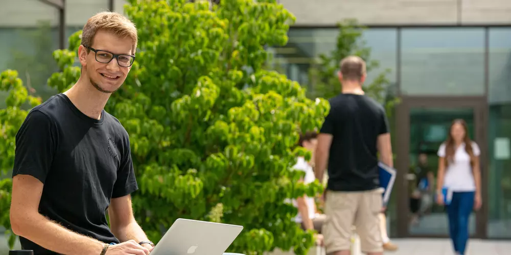  Student sitzt mit Laptop in Innenhof und lernt. Im Hintergrund sieht man weitere Studierende.