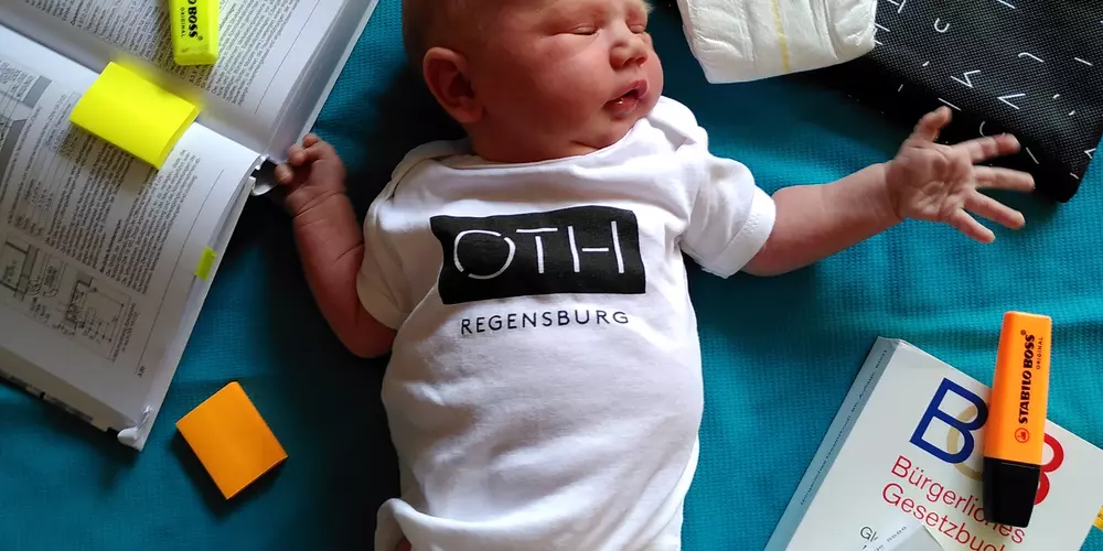 Baby trägt den OTH-Body, liegt  auf einer türkisen Decke zwischen Gesetzestexten, Mitschriften, Textmarkern und einer Windel.