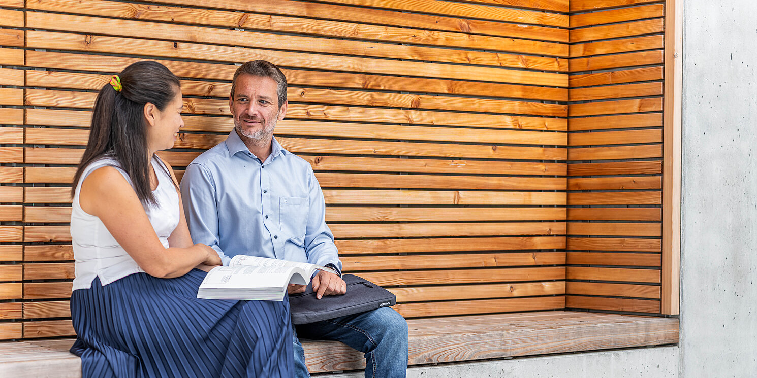 Module aus dem Master Business Administration – Leadership, Management und Transformation berufsbegleitend absolvieren Zwei Studierende sitzen auf einer Bank und unterhalten Sich.