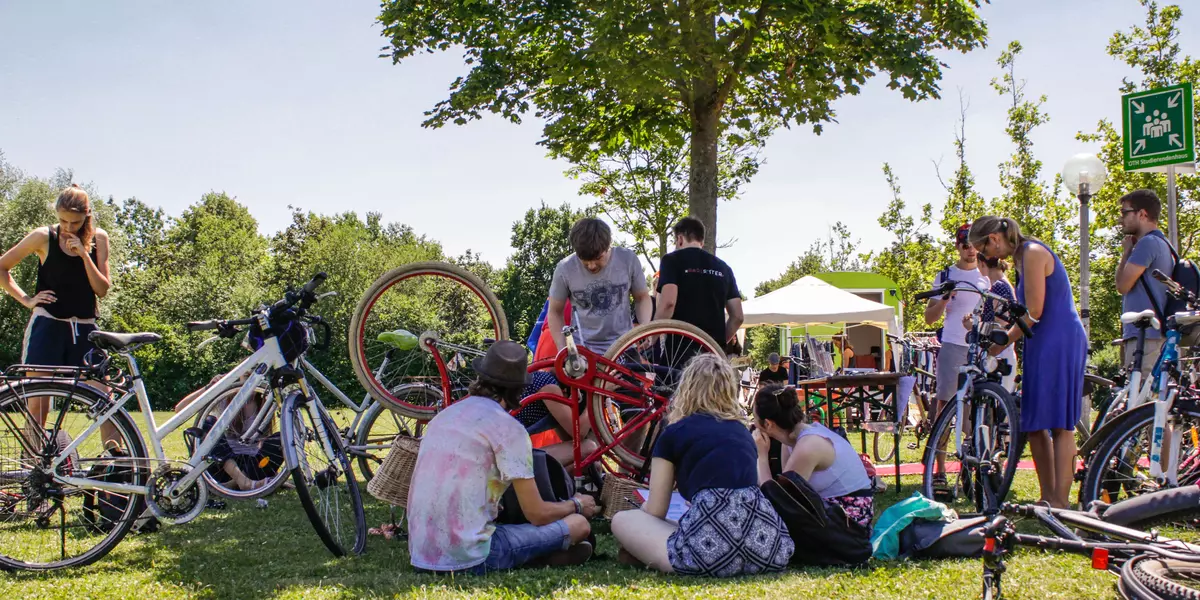 Auf dem Foto ist eine Gruppe von Menschen zu sehen, die gemeinsam auf einer Wiese Fahrräder reparieren. 