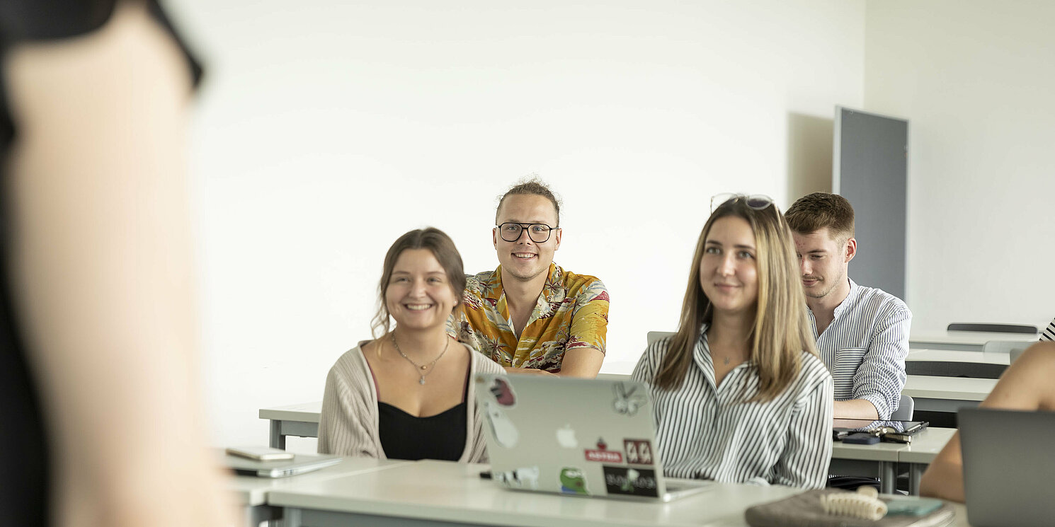 Eine Gruppe von vier Studierenden sitzt in einem Vorlesungssaal, im Vordergrund sieht man die Professorin. 