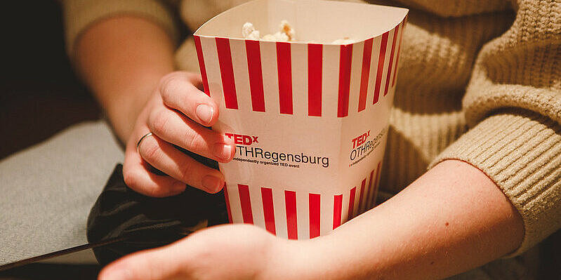 Zwei Hände halten einen Popcorn-Eimer mit der Aufschrift TEDxOTH Regensburg