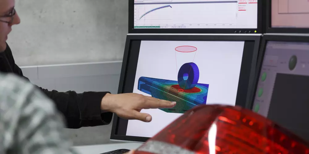 Zwei Männer sitzen an einem Schreibtisch vor einer PKW-Heckleuchte. Ein Bildschirm zeigt die Deformation der Heckleuchte beim Fertigungsschritt des Laserschweißens. Verschiedene Farben verdeutlichen die Bereiche, die aufgrund der Spannungen stark verformt werden.