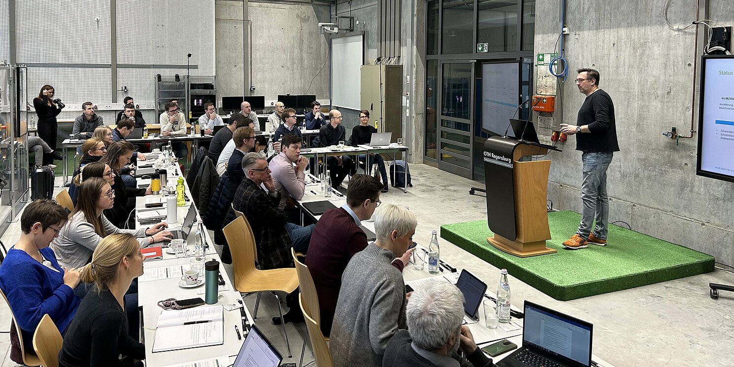 Teilnehmende sitzen in einem Hörsaal. Ein Redner steht am Pult. 