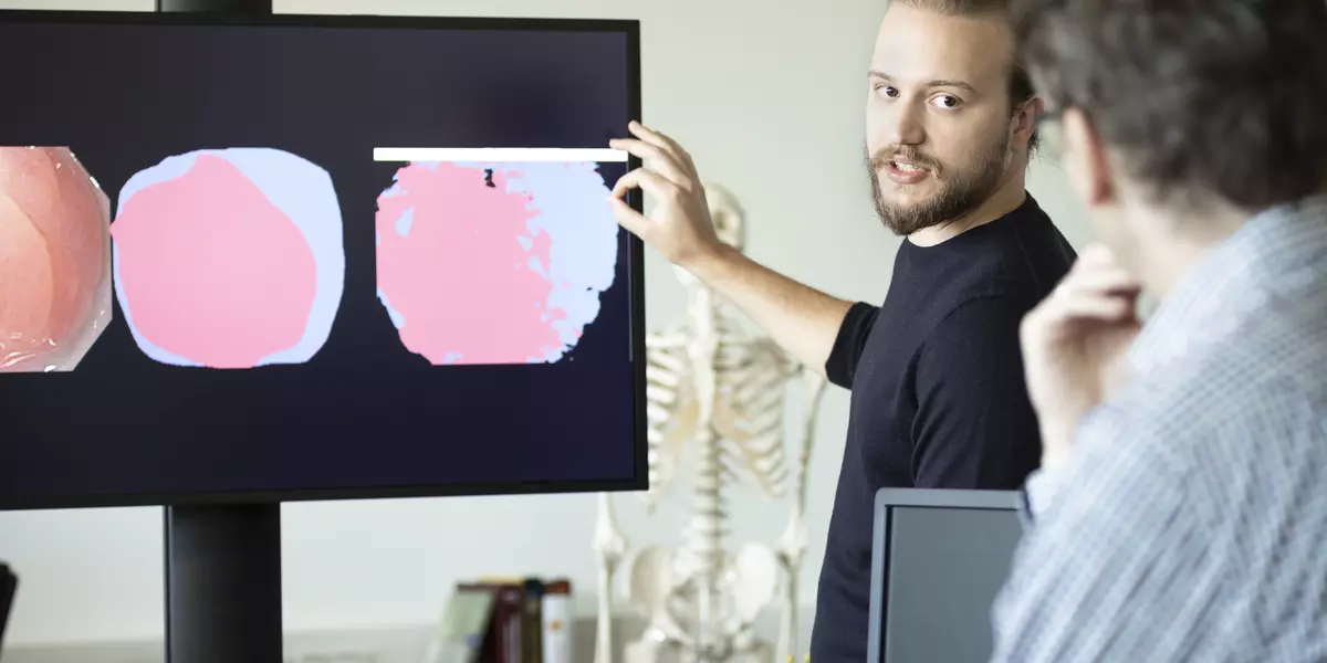 Auf einem Bildschirm sind drei mehrfarbige, kreisförmige Abbildungen abgebildet, die sich voneinander unterscheiden. Ein junger Mann steht vor dem Bildschirm und erläutert, was zu sehen ist.