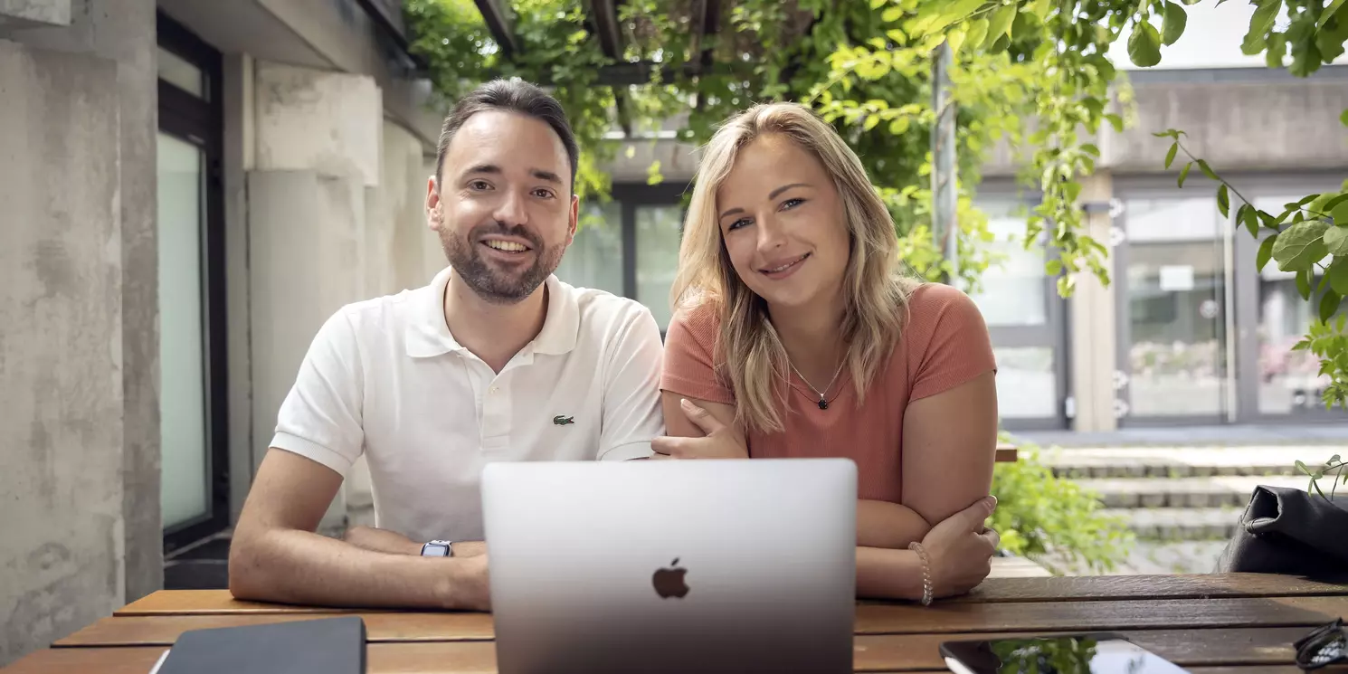 Ein Mann und eine Frau sitzen draußen an einem Tisch vor einem Apple Notebook. Im Hintergrund das OTH Gebäude.