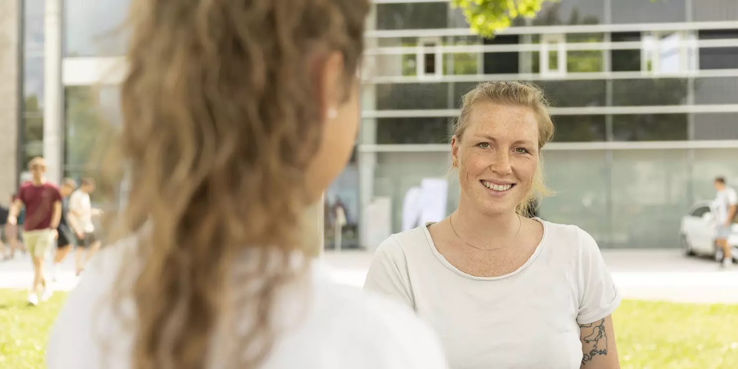 Zwei Frauen stehen sich gegenüber. Eine dunkelhaarige Frau mit Rückenansicht. Eine blonde Frau mit Sommersprossen und weißem T-Shirt.