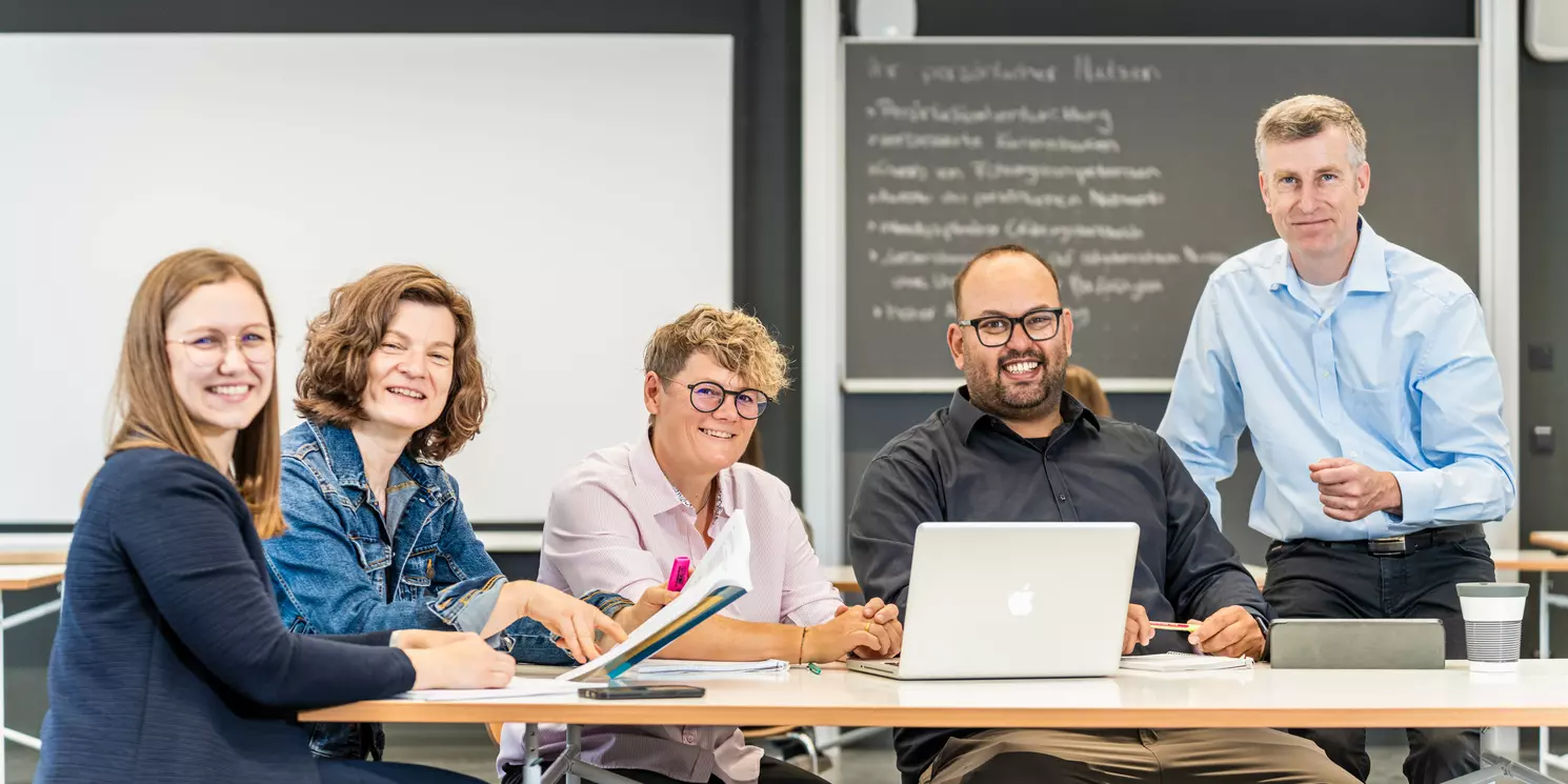 Studierende und ihr Dozent arbeiten mit ihren Lernmaterialien an einem Tisch.