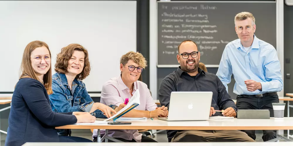 Studierende und ihr Dozent arbeiten mit ihren Lernmaterialien an einem Tisch.