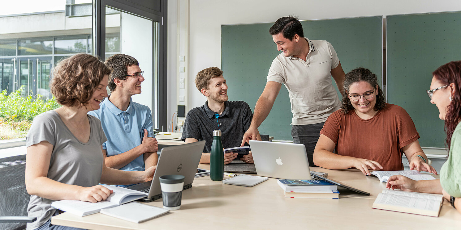 Studierende arbeiten in einer Lerngruppe mit Laptops und Unterlagen an einem großen Tisch.