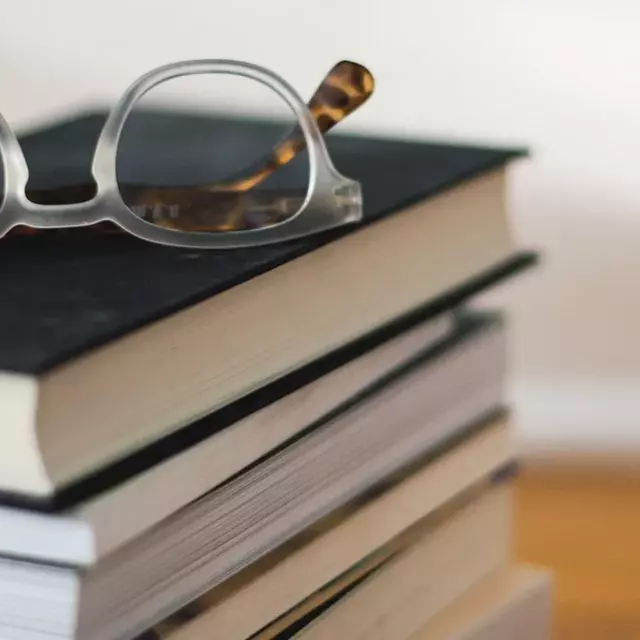Auf einen Holztisch liegt ein Stapel Bücher. Oben drauf liegt eine Brille.