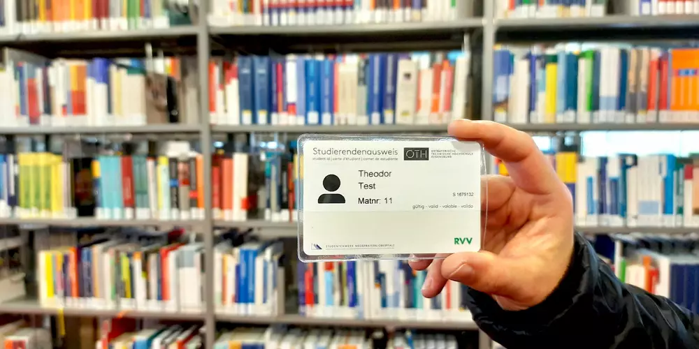 The OTH Regensburg card is held in a hand. The background shows several library books.
