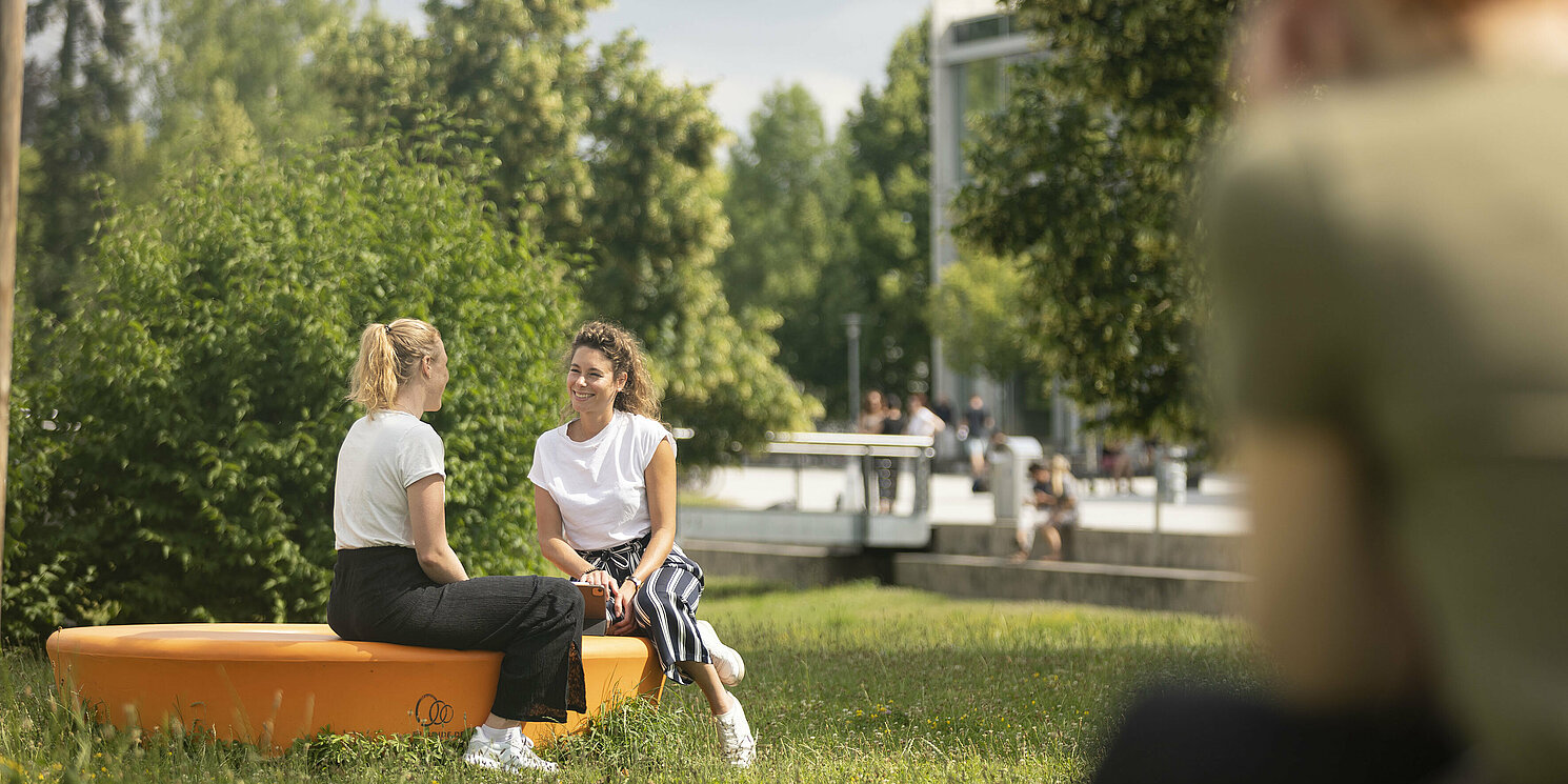 Zwei Frauen sitzen draußen auf den orangen Sitzkreisen. Im Hintergrund Bäume.