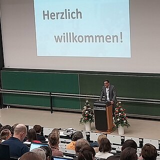 Ein Hörsaal voller Menschen betrachtet den Referenten Prof. Dr. Oliver Steffens. Eine Begrüßungs-PowerPoint wird an die Wand projiziert.