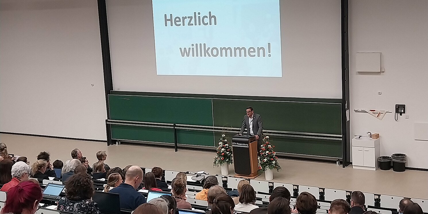 Ein Hörsaal voller Menschen betrachtet den Referenten Prof. Dr. Oliver Steffens. Eine Begrüßungs-PowerPoint wird an die Wand projiziert.