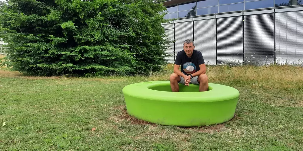 Klaus Kagerer sitzt auf einem grünen Sitzkreis in der Wiese. Im Hintergund das OTH Gebäude