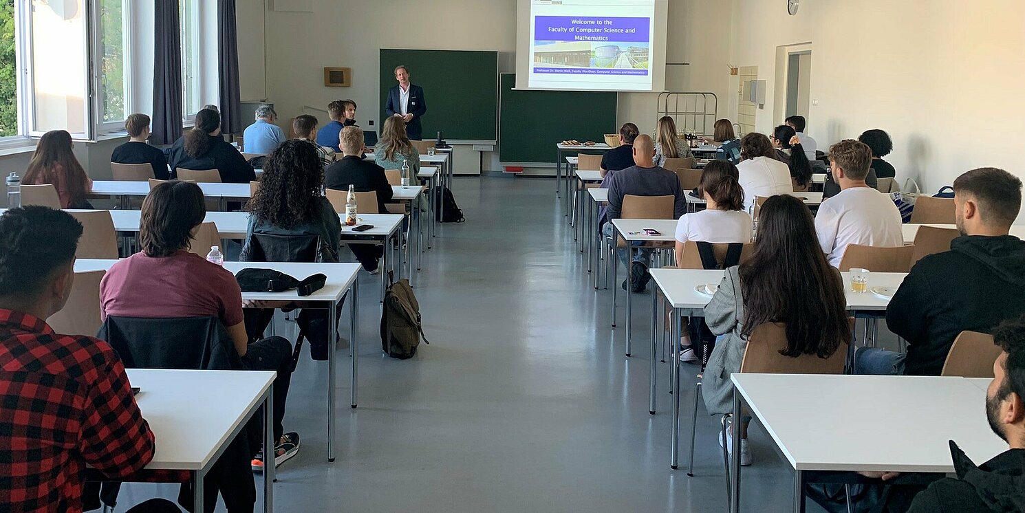 Studierende sitzen in einem Vorlesungsraum. Ein Dozent steht vorne.