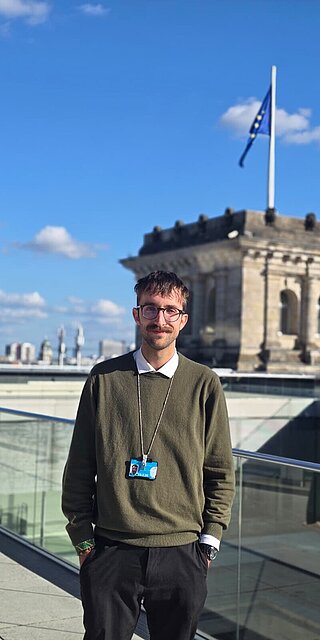 Das Bild zeigt Michael Scharpf vor dem Bundestag in Berlin. Er trägt einen dunkelgrünen Pullover mit darunter liegendem Hemd und hat einen Ausweis umhängen. 