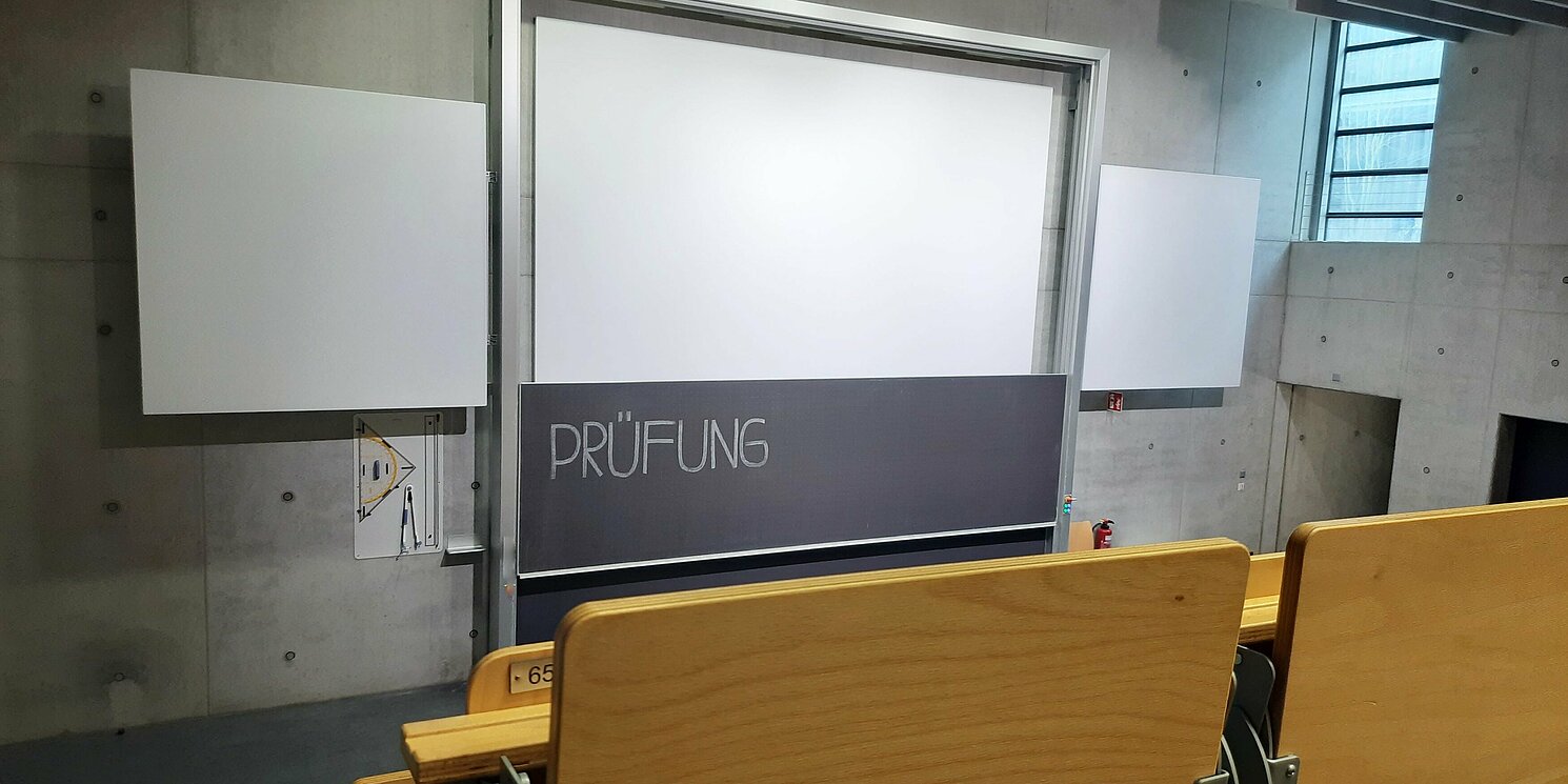 Ein leerer Hörsaal. An der Tafel steht das Wort Prüfung.