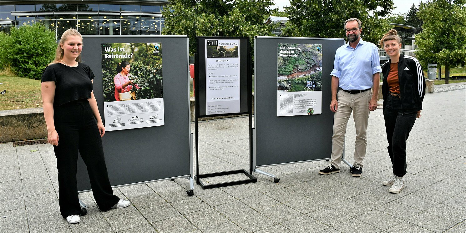 Zwei Frauen und ein Mann stehen neben Plakaten zum Thema Fairtrade und Klimawandel.