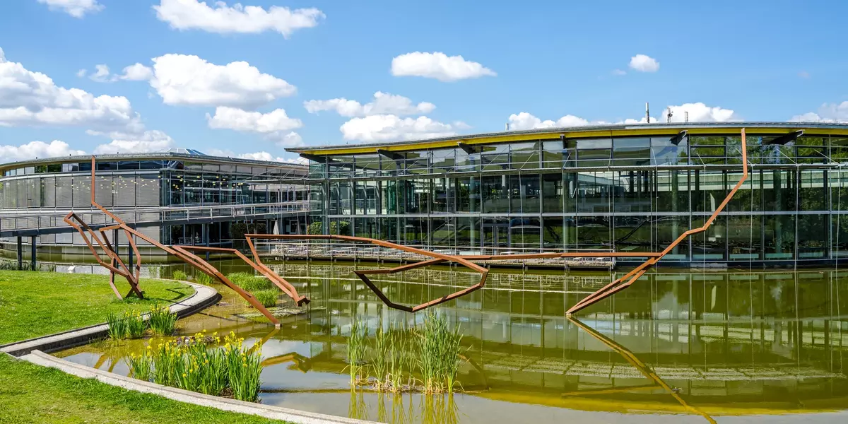 Ein Blick auf den Campus, im Vordergrund Wiese und See mit einer Skulptur von Robert Schad, im Hintergrund die Gebäude von Bibliothek und Mensa, darüber blauer Himmel