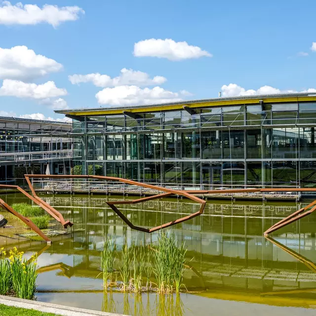 Ein Blick auf den Campus, im Vordergrund Wiese und See mit einer Skulptur von Robert Schad, im Hintergrund die Gebäude von Bibliothek und Mensa, darüber blauer Himmel