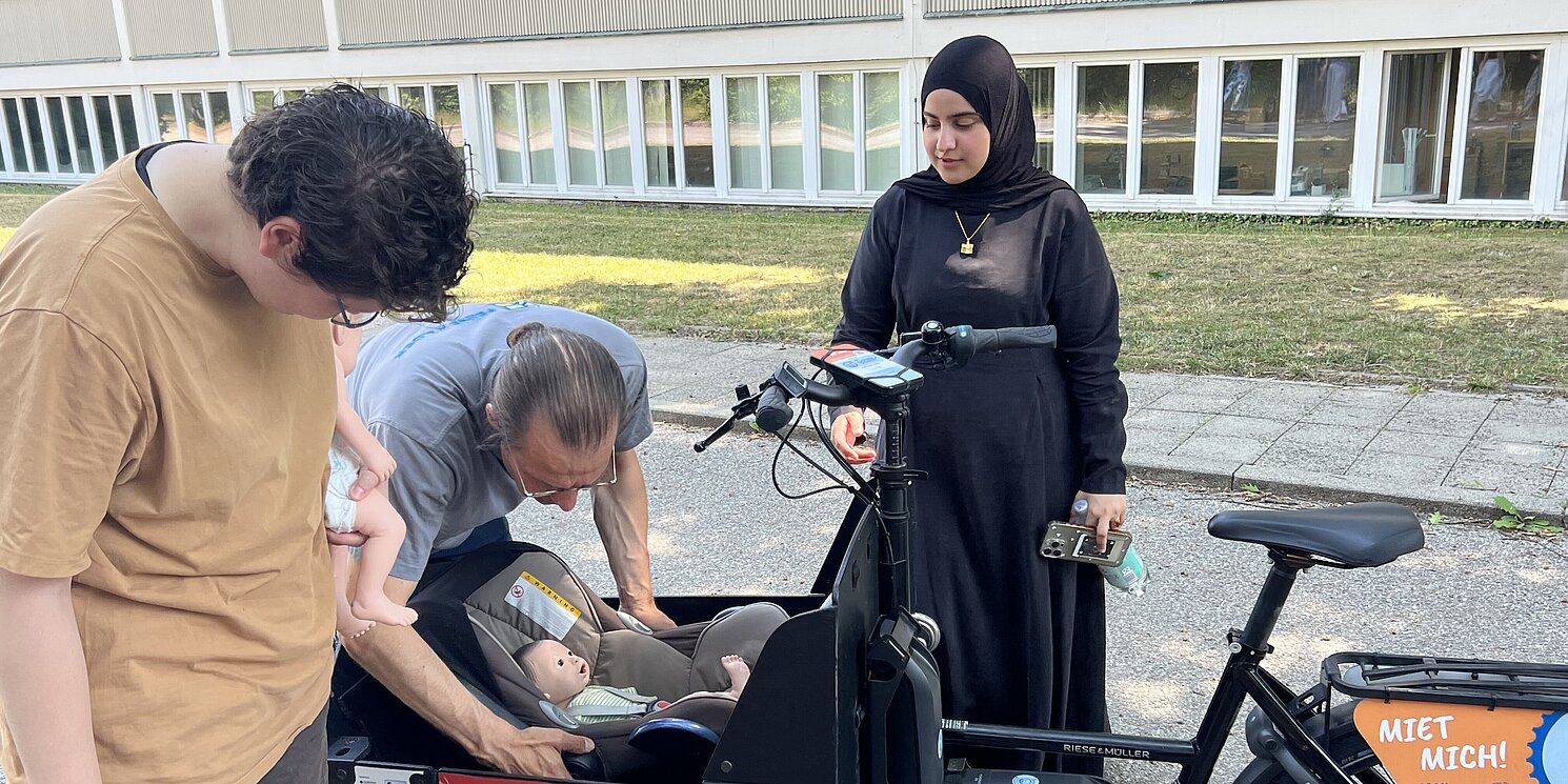 Ein Lastenfahrrad mit einem Korb vorne, in dem man einen Säugling als Puppe gelegt hat. Ein Mitarbeiter von Feine Räder platziert es richtig im korb. Zwei Studenten stehen links und rechts daneben und sehen es die korrekte Anbringung an.