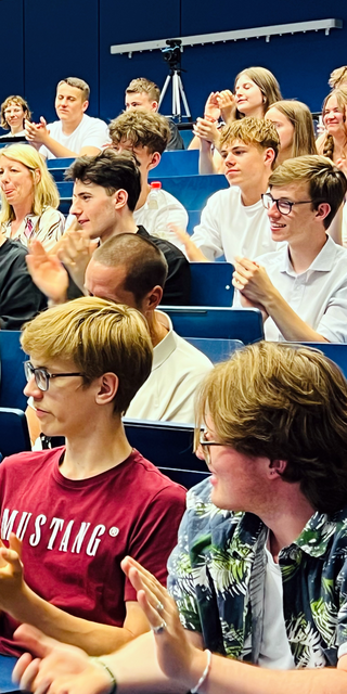 Ein voller Hörsaal, die Schüler klatschen und nehmen ausgelassen an der Feier teil. 