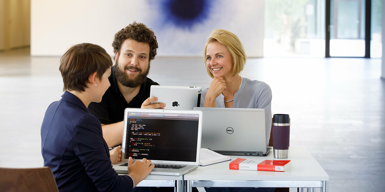 Drei Studierende sitzen in einer Lerngruppe mit Laptops und Büchern zusammen