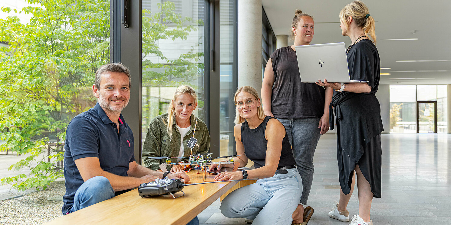 Studierende sitzen und stehen um eine Bank, auf der ein Roboter steht und arbeiten mit ihm.