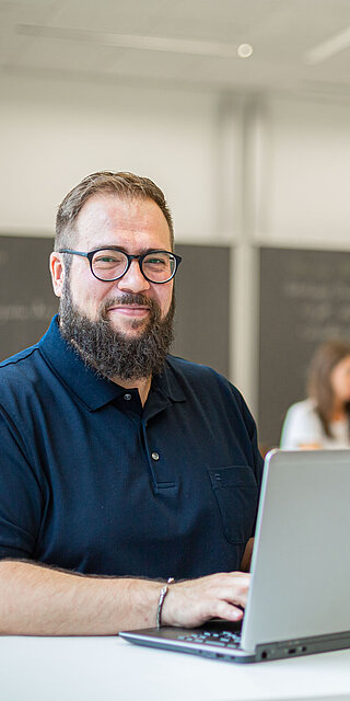 Ein Mann mittleren Alters sitzt an seinem Laptop in einem Seminarraum.