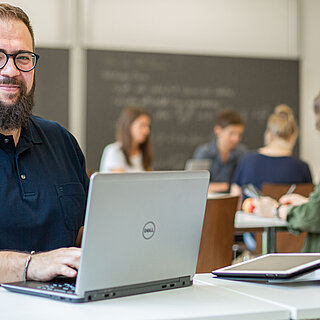 Ein Mann mittleren Alters sitzt an seinem Laptop in einem Seminarraum.