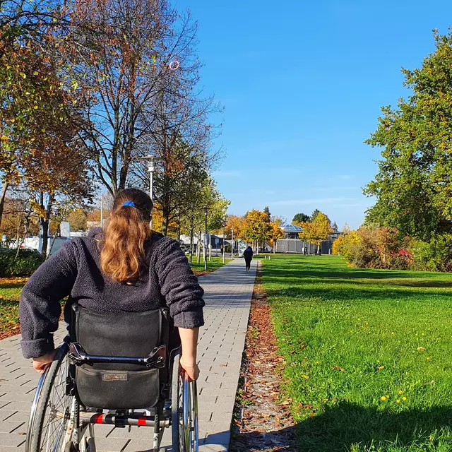 Eine Rollstuhlfahrerin auf einem Weg am OTH Campus. Rechts und links sind Bäume und eine grüne Wiese.