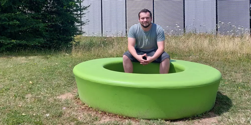 Florian Moosbauer sitzt auf einem grünen Sitzkreis in der Wiese. Im Hintergund das OTH Gebäude