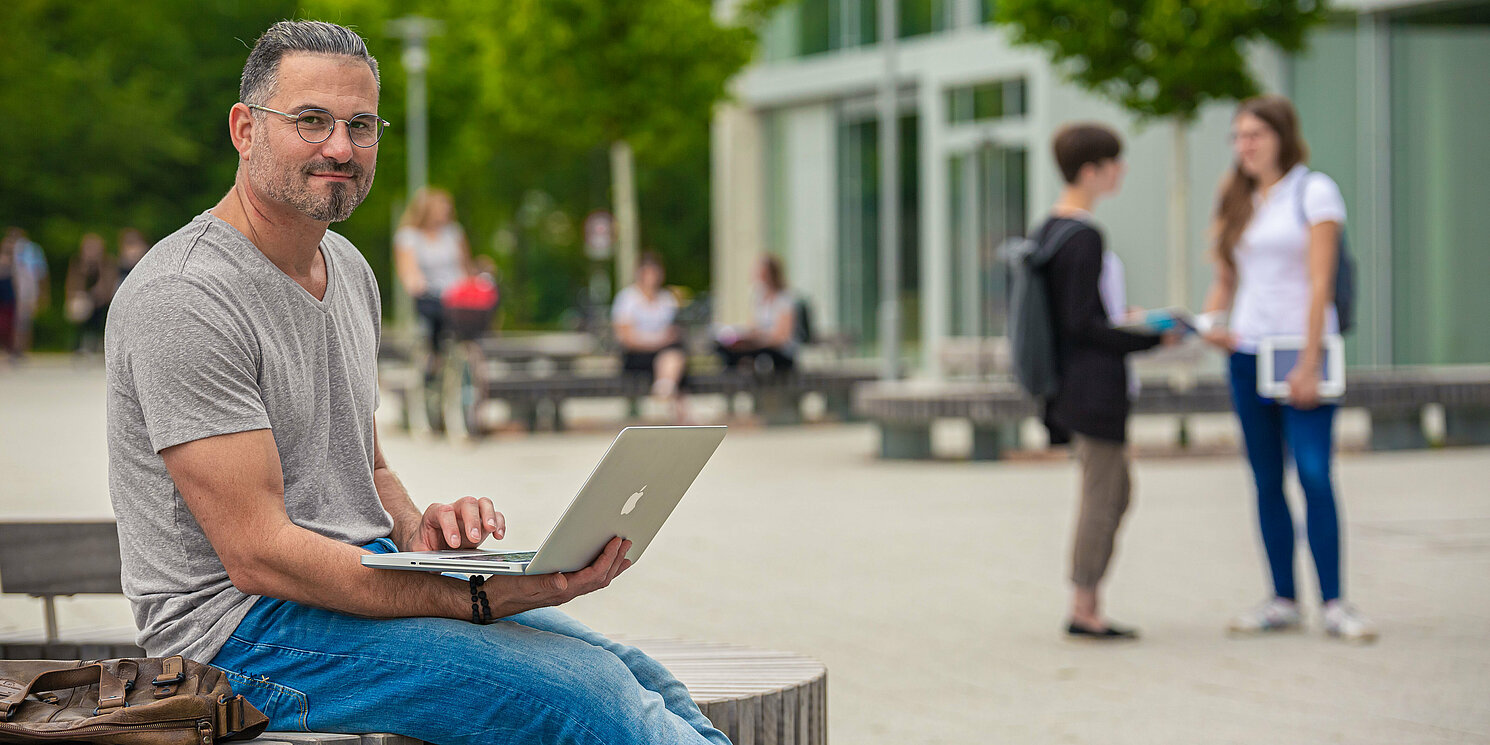 Student im Freien mit Laptop auf einer Bank. Im Hintergrund sieht man weitere Studierende auf dem Campus.