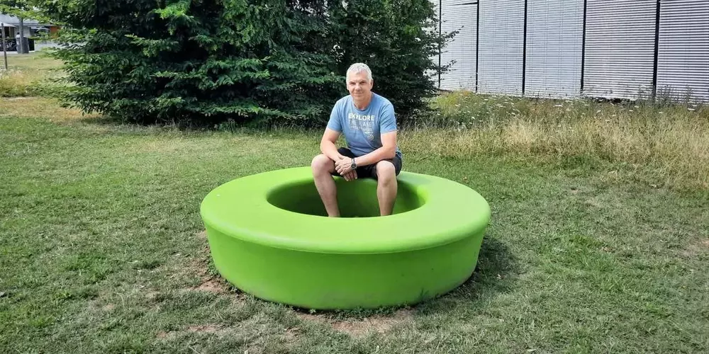 Robert Deibl sitzt auf einem grünen Sitzkreis in der Wiese. Im Hintergund das OTH Gebäude