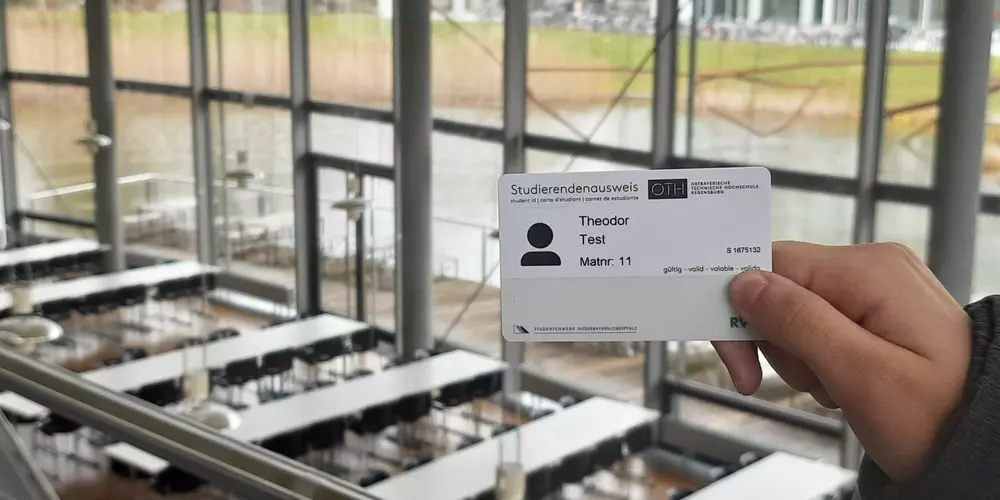 The OTH Regensburg card is held in a hand. The background shows some of the cafeteria tables, the small pond in front of the building and a small part of the Haus der Technik.