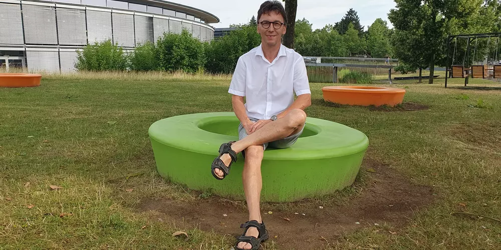 Franz Wagner sitzt auf einem grünen Sitzkreis in der Wiese. Im Hintergund das OTH Gebäude
