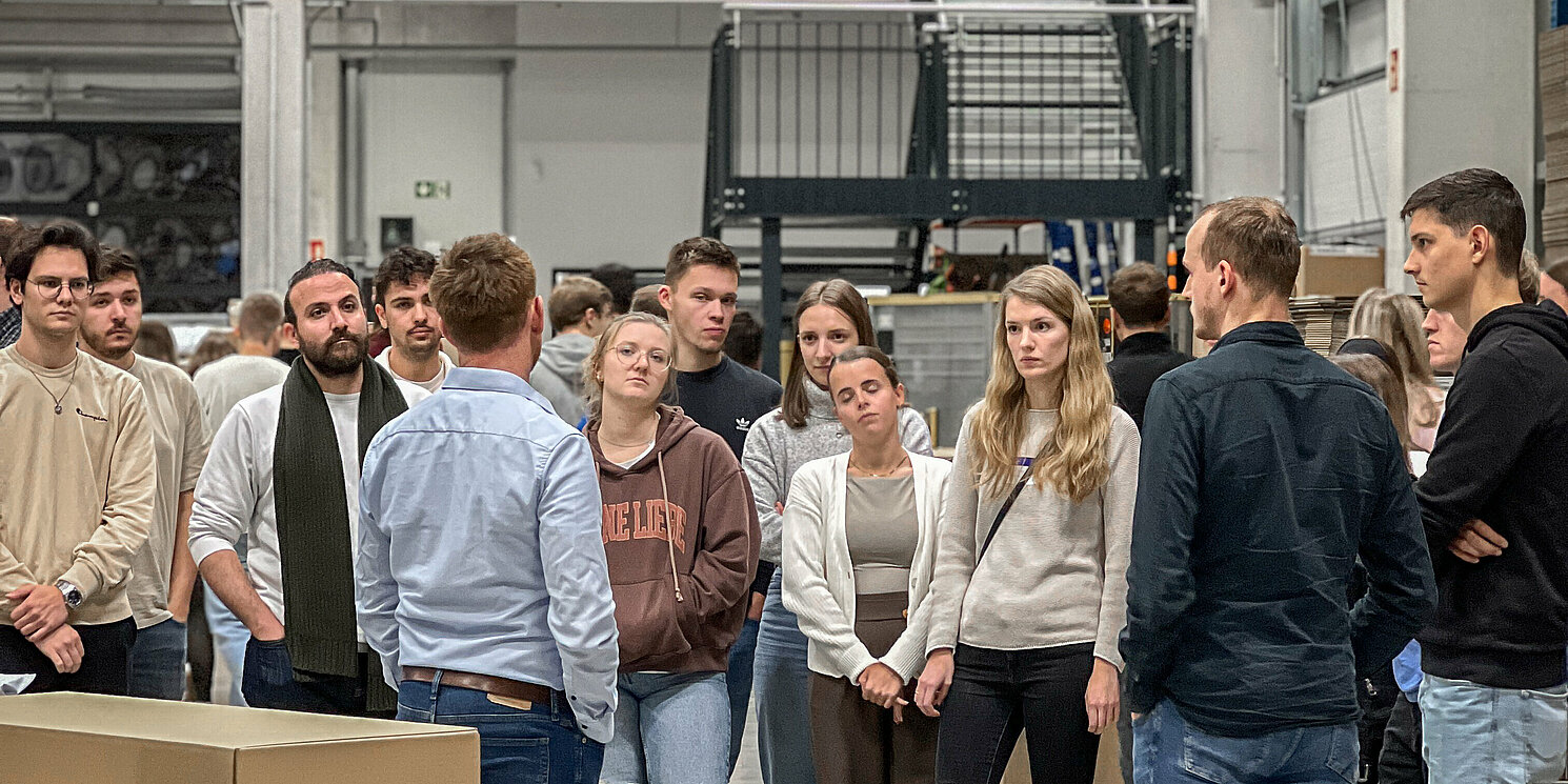 Eine Gruppe von Studierenden stehen in einem Raum, im Hintergrund Pakete und Regale