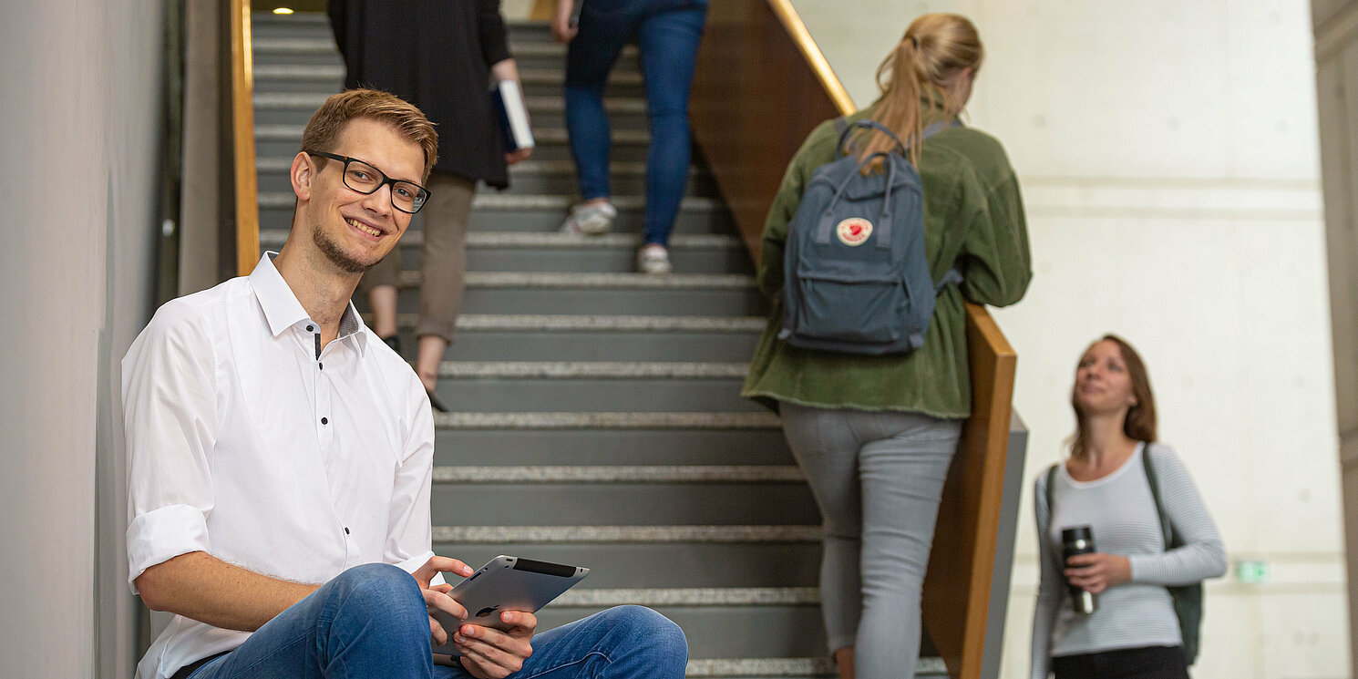 Student sitzt mit Tablet auf einer Treppe. Im Hintergrund sind weitere Studierende zu sehen. 
